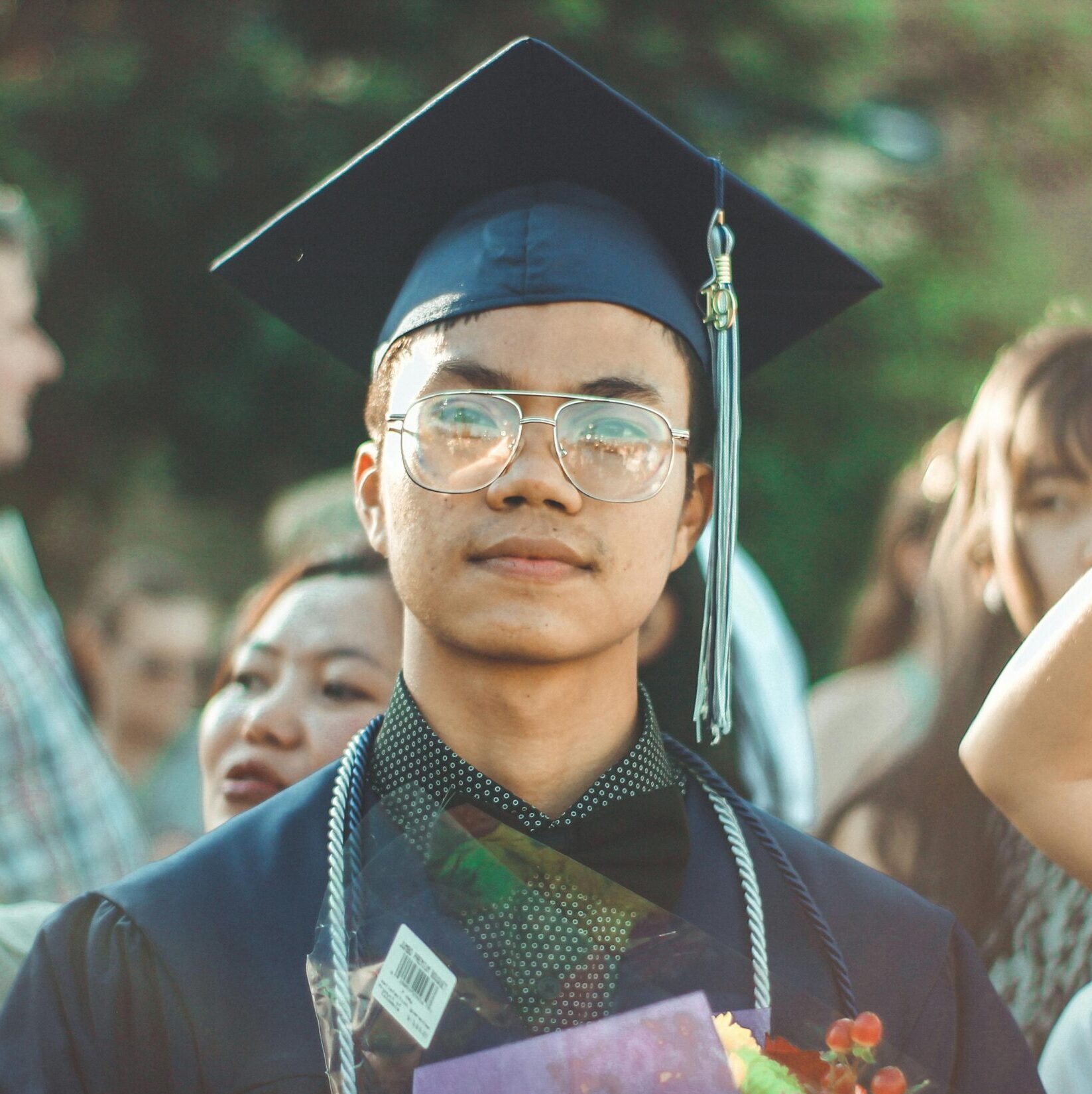 Asian male graduate smiling during an outdoor graduation celebration with family and friends.