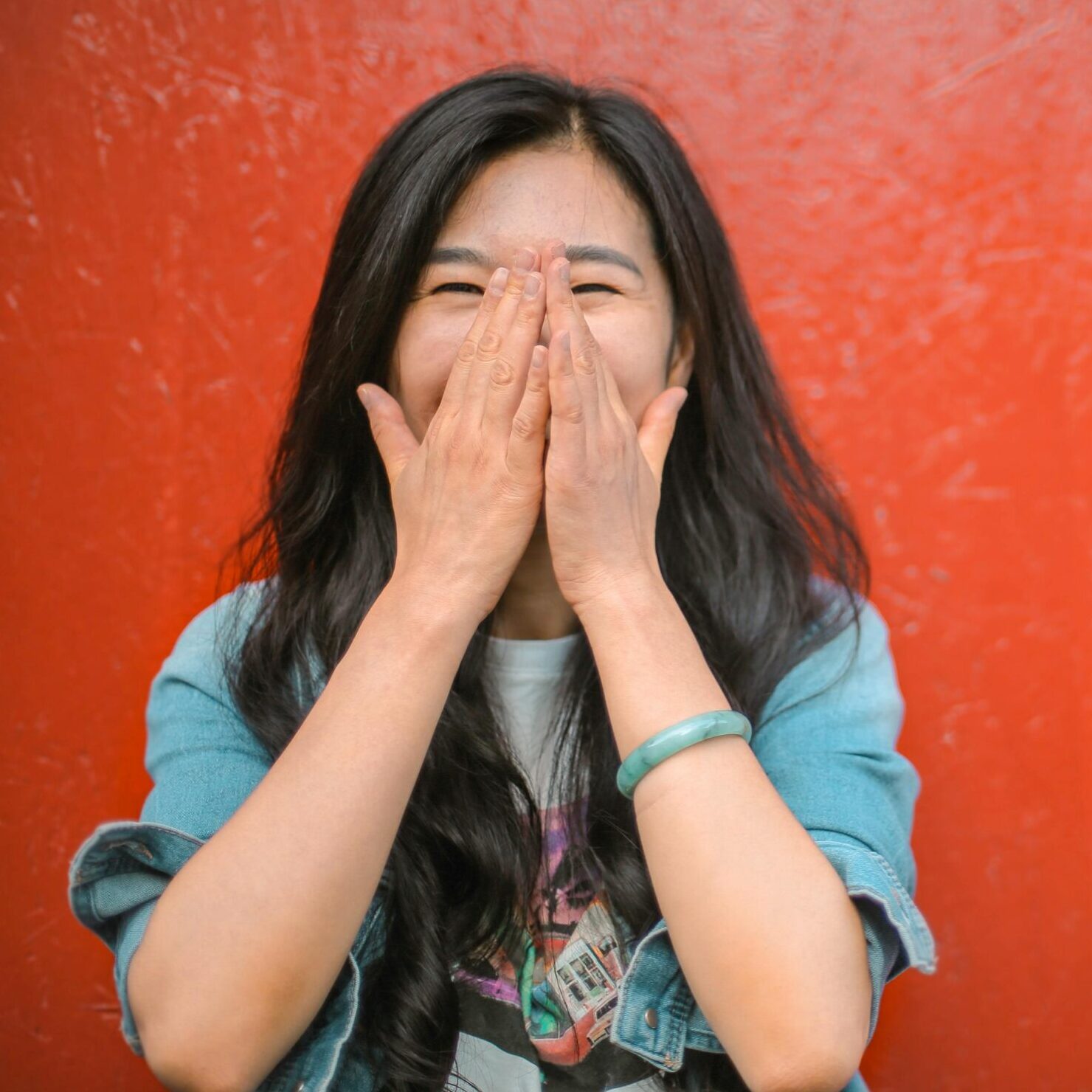 Young overjoyed Asian female in casual outfit covering face with hands and laughing while standing against vibrant orange wall